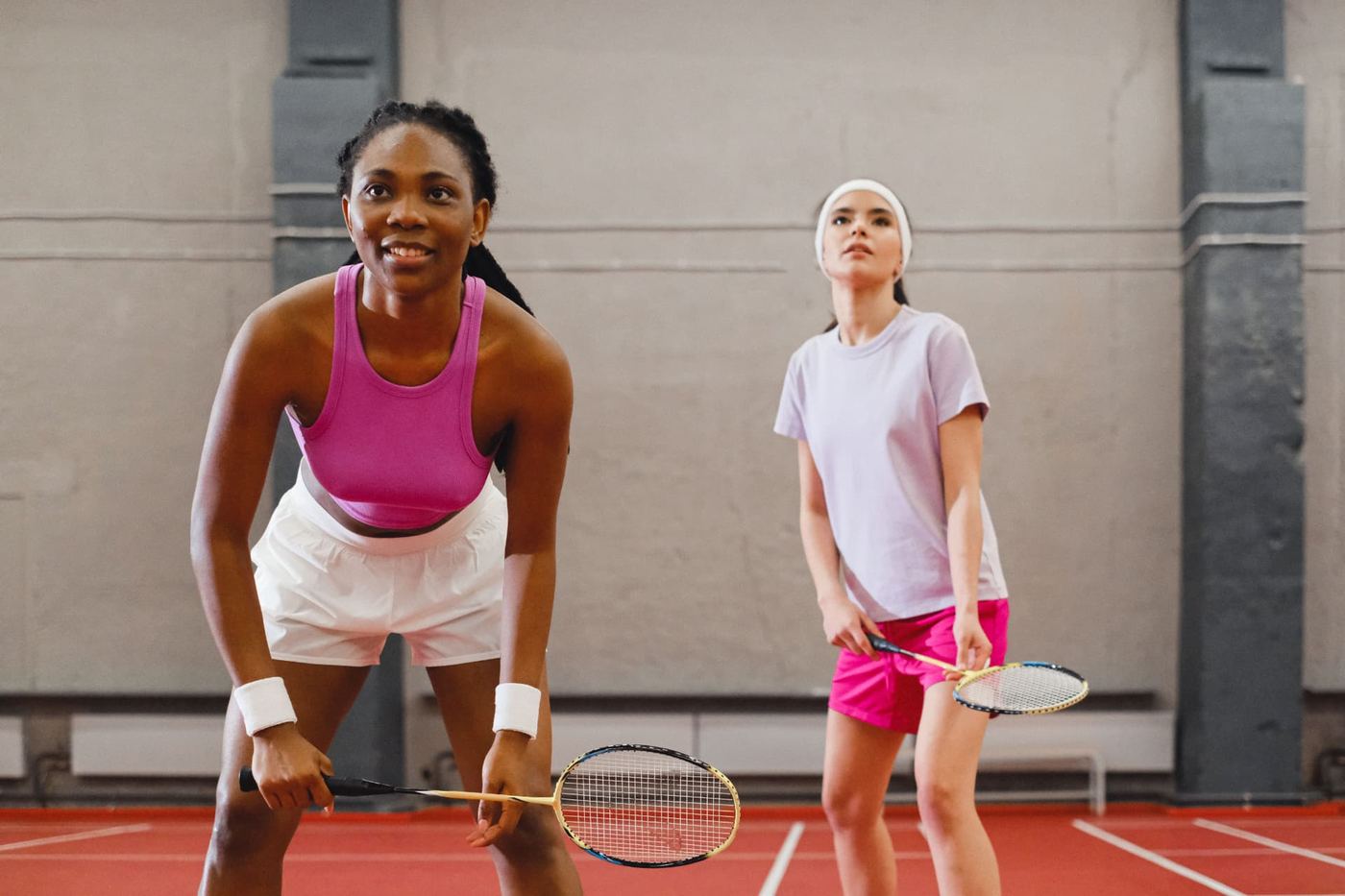 Two badminton players crouch in a ready stance with racquets low on a red indoor court.